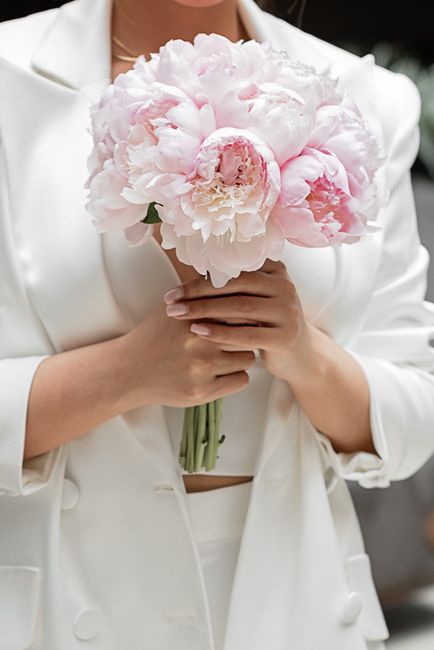 A person dressed in a white blazer is holding a bouquet of large, soft pink peonies with lush, ruffled petals and green stems. The bouquet is tightly arranged and appears fresh with a delicate texture