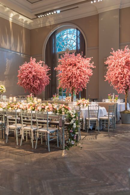 An elegant wedding reception setup featuring long banquet tables decorated with floral garlands in shades of pink, white, and blush, complemented by silver Chiavari chairs. Three large, lush pink cher