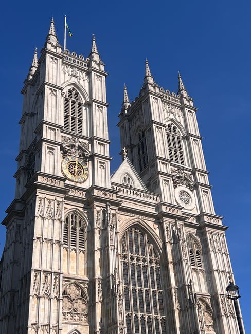 The image shows the exterior of Westminster Abbey, a historic Gothic church with two tall, ornate towers crowned with spires, set against a clear blue sky. The facade features large arched windows wit