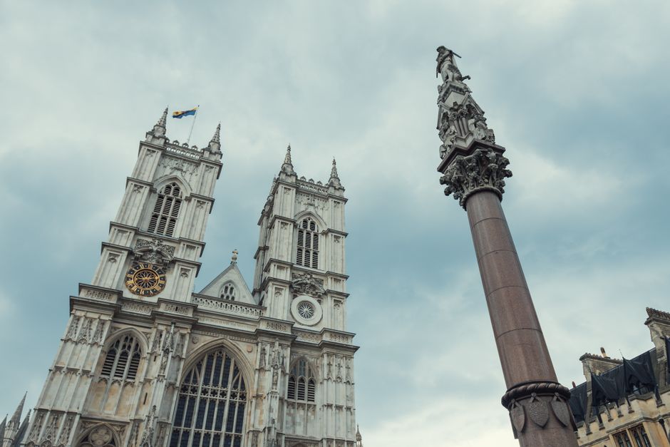 A detailed view of Westminster Abbey's Gothic façade featuring two prominent spires adorned with ornate stone carvings, large arched windows, and a clock with gold accents set against a cloudy sky. T