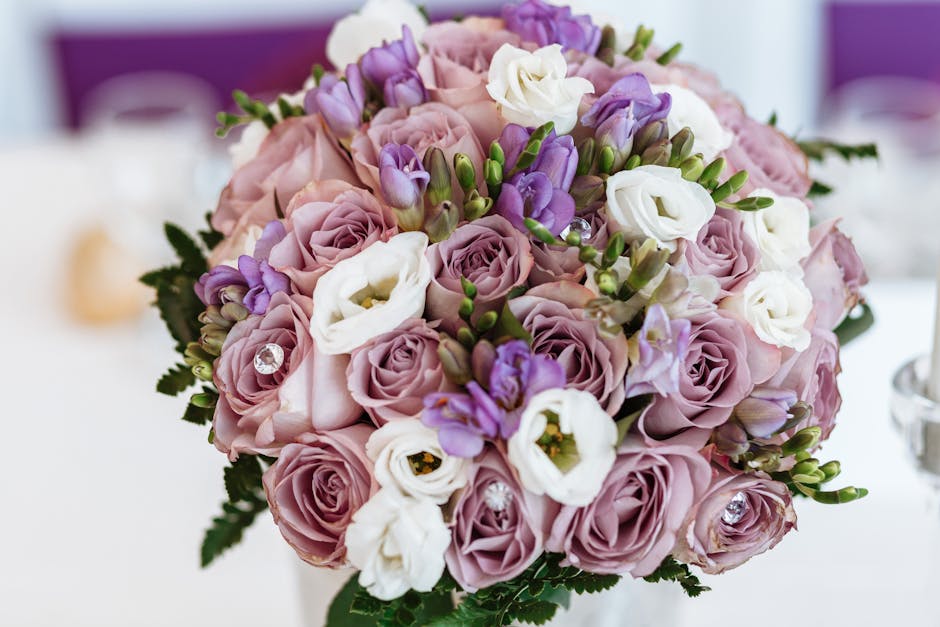 A close-up view of a floral bouquet arranged with soft pink roses, white eustoma, and purple freesias, accented with green foliage and small decorative rhinestones. The flowers are freshly cut, with p