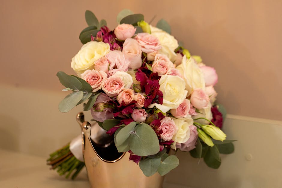 A floral bouquet featuring a mix of fresh white roses, soft pink spray roses, and deep burgundy blooms arranged with eucalyptus leaves. The bouquet is placed in a metallic vase or container, with the 